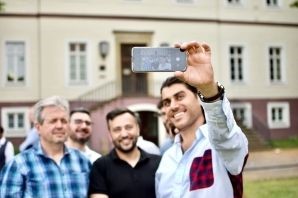 Vier Männer stehen vor einem Haus und schauen auf ein Smartphone, das einer der Männer in der Hand hält. Vier Männer stehen vor einem Haus und schauen auf ein Smartphone, das einer der Männer in der Hand hält.