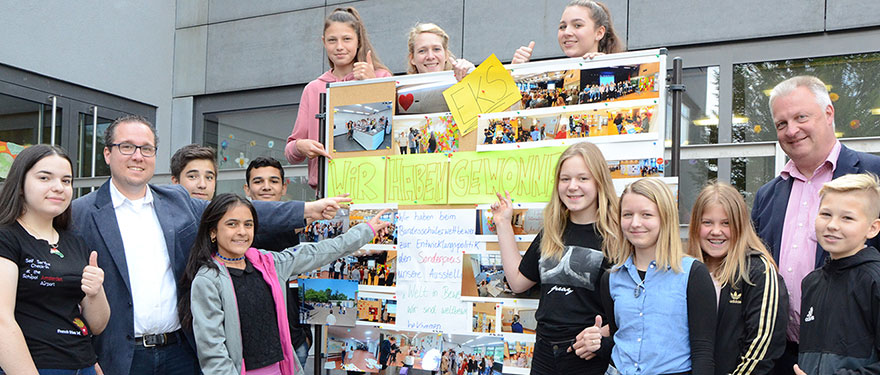 Schüler und Lehrer stehen an einer Stellwand auf der Fotos und der Schriftzug steht: "Wir haben gewonnen".