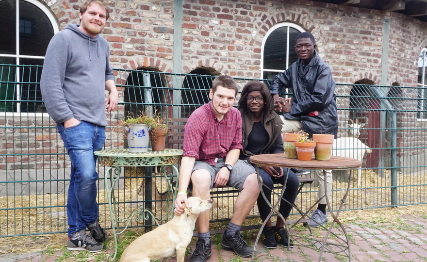 Zwei deutsche Gastgeber und zwei ghanaische Gäste sitzen und stehen an einem Tisch vor einem Gebäude des Biohofs, vor ihnen sitzt ein Hund, den einer der Gastgeber streichelt.