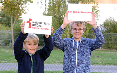 Zwei Jungen halten Schildern mit dem Logo und dem Claim des Bistums in die Luft. Zwei Jungen halten Schildern mit dem Logo und dem Claim des Bistums in die Luft.