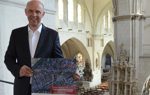 Dompropst Kurt Schulte steht auf einer Balustrade mit dem Inneren des Doms im Hintergrund und hält ein Plakat der DomGedanken in den Händen.