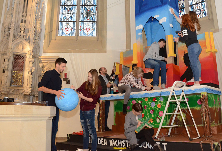 Pastoralreferent Jens König-Upmeyer (links) hält den Globus, den Jacqueline Kijowski gestaltet. Im Hintergrund bemalen Schülerinnen sowie Schüler aus Religions- und Kunstkursen das Holzmodell bunt.