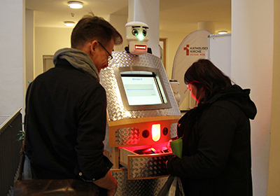 Zwei Personen lassen sich von einem Segensroboter segnen. Zwei Personen lassen sich von einem Segensroboter segnen.