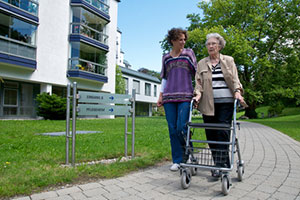 Eine junge Frau begleitet eine Seniorin mit Rollator.