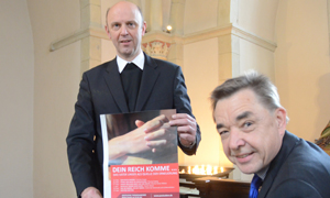 Dompropst Kurt Schulte, stehend, hält ein Plakat der Veranstaltungsreihe "Geistliche Themenabende" in den Händen. Rechts vor ihm sitzt Thomas Söding.