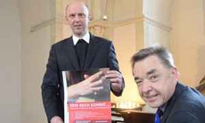 Dompropst Kurt Schulte, stehend, hält ein Plakat der Veranstaltungsreihe "Geistliche Themenabende" in den Händen. Rechts vor ihm sitzt Thomas Söding. Dompropst Kurt Schulte, stehend, hält ein Plakat der Veranstaltungsreihe "Geistliche Themenabende" in den Händen. Rechts vor ihm sitzt Thomas Söding.