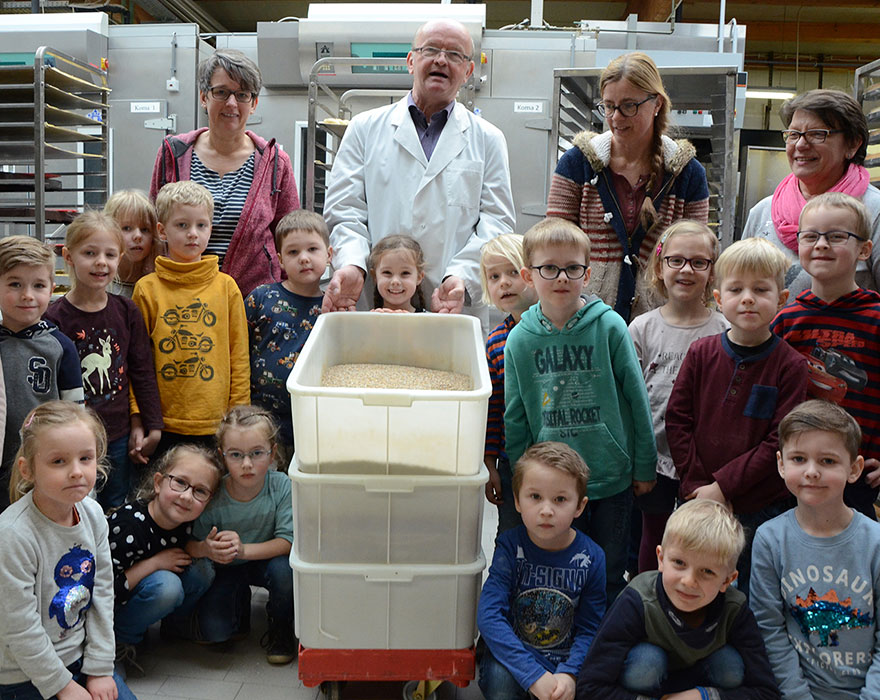 Die Kinder stehen gemeinsam mit ihren Erzieherinnen und dem Bäcker Willi Ebbing um Kisten mit einer Körnermischung herum.