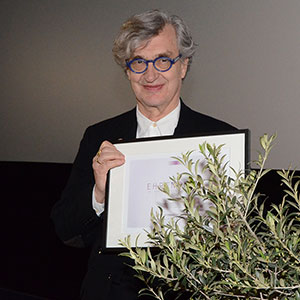 Wim Wenders hält die Urkunde in der Hand und steht hinter dem Olivenbaum.