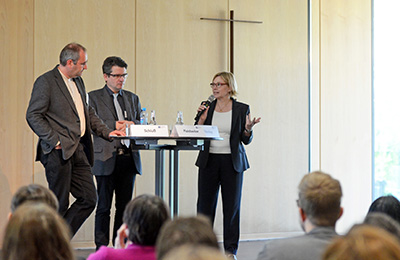 Drei Personen diskutieren auf einem Podium miteinander. Drei Personen diskutieren auf einem Podium miteinander.