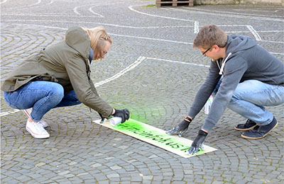 Zwei Personen sprayen mit einer Schablone den Schriftzug "#meinfokus19" auf die Straße. 