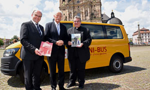 Vor einem gelben Kleinbus, einem BONI-Bus, präsentieren (von links) Martin Guntermann, Heinz Paus und Monsignore Georg Austen den Jahresbericht. Im Hintergrund ist der Dom zu Fulda zu sehen.