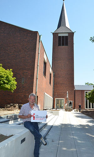 Michael Oetterer sitzt vor der umgebauten Kirche und hält ein Schild in der Hand mit der Information "Familienbildungsstätte Dorsten-Marl".