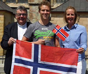 Vor einer Kirchenfassade stehen (von links) Georg Austen, Lasse Haack und Julia Jesse. Die beiden Außenstehenden halten gemeinsam an beiden Seiten fassend eine große norwegische Flagge vor die Gruppe, Lasse Haack ein kleines Fähnchen.