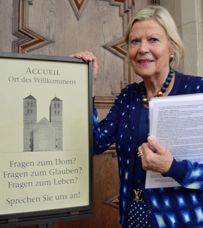 Links ein Schild mit der Aufschrift "Accueil - Ort des Willkommens. Fragen zum Dom? Fragen zum Glauben? Fragen zum Leben? Sprechen Sie uns an!" Links vom Schild steht Helga-Dietlind Rahn mit Unterlagen unter dem linken Arm, die rechte Hand auf das Schild gelegt.