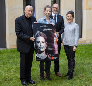 Vor einer Wand mit Fenstern stehen (von links): Felix Genn, Sabrina Friedrich, Diethelm Schaden und Simone Simons. Genn und Friedrich halten gemeinsam das Plakatmotiv, das Sabrina Friedrichs Gesicht neben einem strahlenden Kirchenfenster vor schwarzem Hintergrund abbildet. Darauf steht zu lesen: "Glaube braucht Räume und tatkräftige Bauingenieure (m/w/d)."			