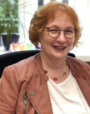 Maria Bubenitschek, mit kinnlangen roten Haaren und Brille und gekleidet in eine cognacfarbene Lederjacke mit cremefarbenem Shirt darunter, sitzt lächelnd vor einem Fenster und einer Grünpflanze im Hintergrund.