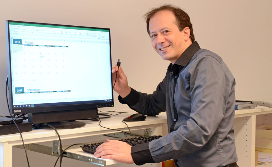 Dimitrios Gieske sitzt neben einem Bildschirm, schaut lächelnd in die Kamera und hält das Ende eines Kabels in der Hand.