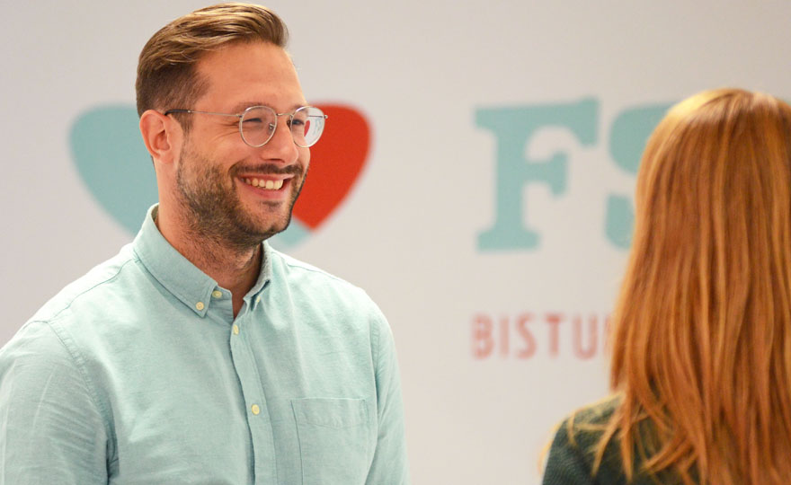 Alexander Oldiges, ein junger Mann mit braunen Haaren, kurzem Bart und Brille, steht links im Bild und unterhält sich mit einer Frau, von der nur der Hinterkopf mit den langen Haaren zu sehen ist. Im Hintergrund ist das Logo der Freiwilligendienste im Bistum Münster zu erkennen.