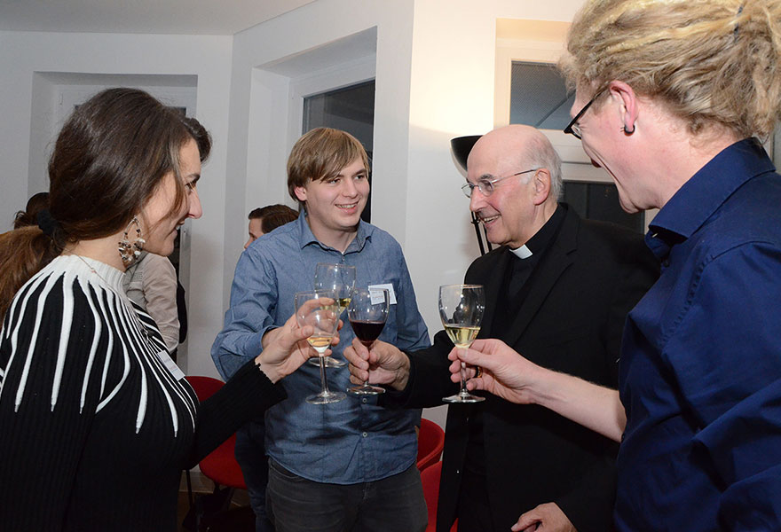 Der Bischof steht mit jungen Erwachsenen zusammen und stößt mit ihnen mit einem Glas Wein an.