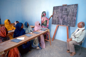 Eine afrikanische Frau schreibt an einer Wandtafel. Links von ihr sitzen weitere afrikanische Frauen in Schulbänken. Rechts sitzt ein Missionar auf einem Hocker. Alle blicken auf die Tafel.