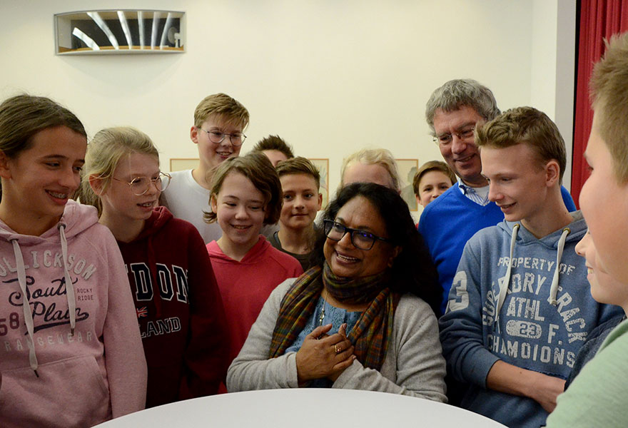 Rita Panicker steht umringt von Schülerinnen und Schülern an einem Stehtisch und freut sich.