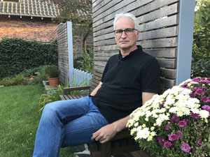 Martin Schmitz sitzt mit Jeans und Pullover bekleidet in einem Garten auf einer Bank Martin Schmitz sitzt mit Jeans und Pullover bekleidet in einem Garten auf einer Bank