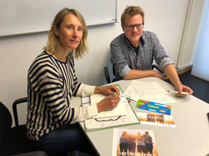 Stefanie Uphues und Christoph Aperdannier sitzen nebeneinander an einem kleinen Tisch. Vor ihnen liegen bunte Faltblätter und Notizzettel.