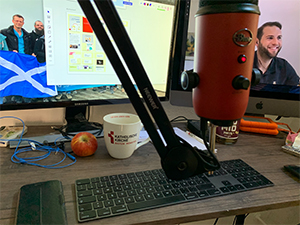 Ein Schreibtisch , auf dem eine Tastatur, eine Tasse und ein Apfel liegen. Im Vordergrund ein Mikrofon, im Hintergrund zwei Bildschirme, auf denen ein Mann als Teilnehmer einer digitalen Konferenz bzw. ein Aufgabenkalender zu sehen sind.