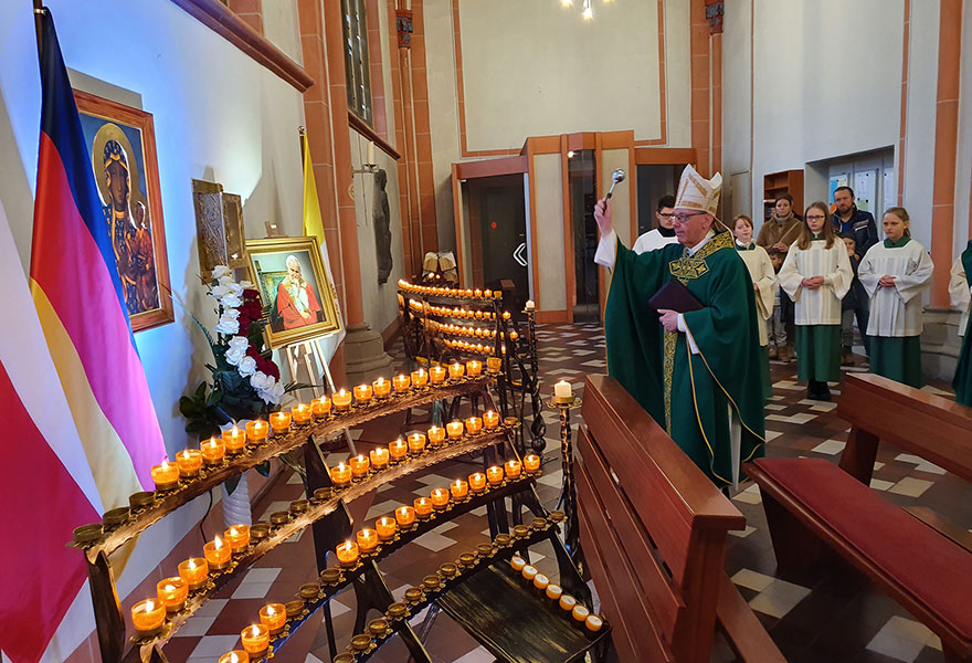 Weihbischof Geerlings segnet den Schrein, der in eine Wand eingelassen ist. Viele Kerzen sind vor dem Gedenkort mit einer Ikone und einem Bild von Johnnes Paul II. angeordnet.