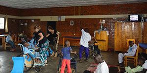 In einem großen Raum sind Menschen mit Behinderung in Rollstühlen zu sehen. Zwei junge Frauen kümmern sich um sie.