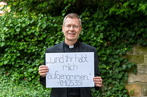 Weihbischof Stefan Zekorn hält ein Plakat mit der Aufschrift "... und ihr habt mich aufgenommen (Mt 25,35)" in den Händen.