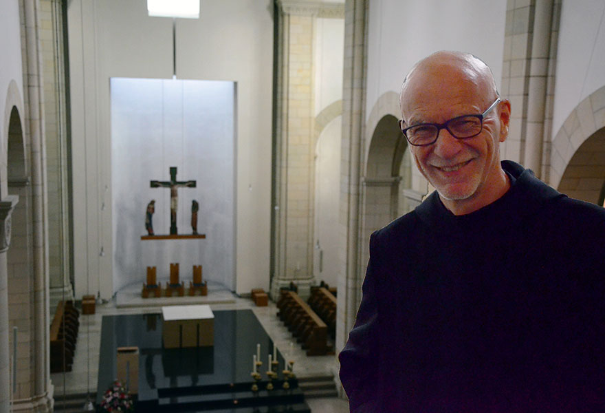 Porträt von Pater Marcel, im Hintergrund sieht man die Abteikirche in Gerleve mit einer Kreuzigungsgruppe.