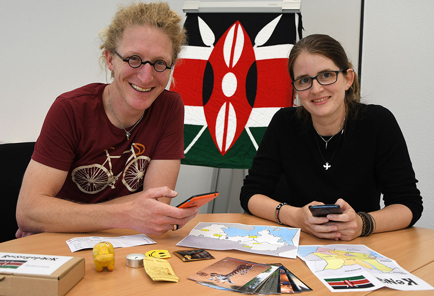 Daniel Gewand und Yvonne Meier sitzen nebeneinander und lächeln in die Kamera. Auf dem Tisch liegt unter anderem eine Landkarte und im Hintergrund ist ein Tuch mit den Farben Kenias aufgehängt.