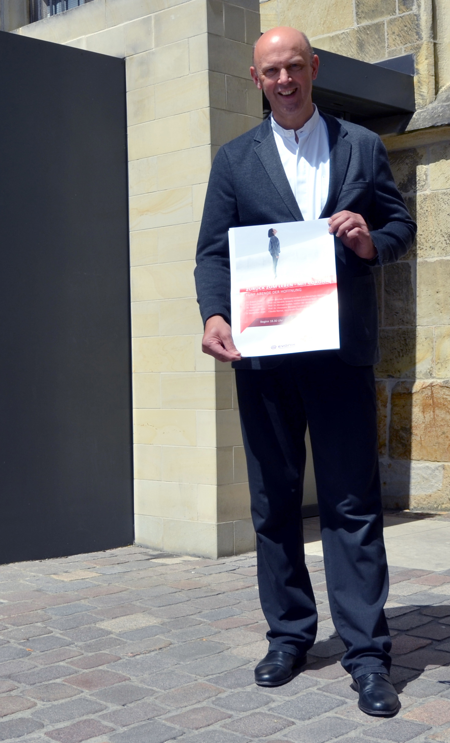 Dompropst Kurt Schulte steht mit einem Plakat der DomGedanken 2020 vor einem Seiteneingang des Doms, dem Uhrenportal.