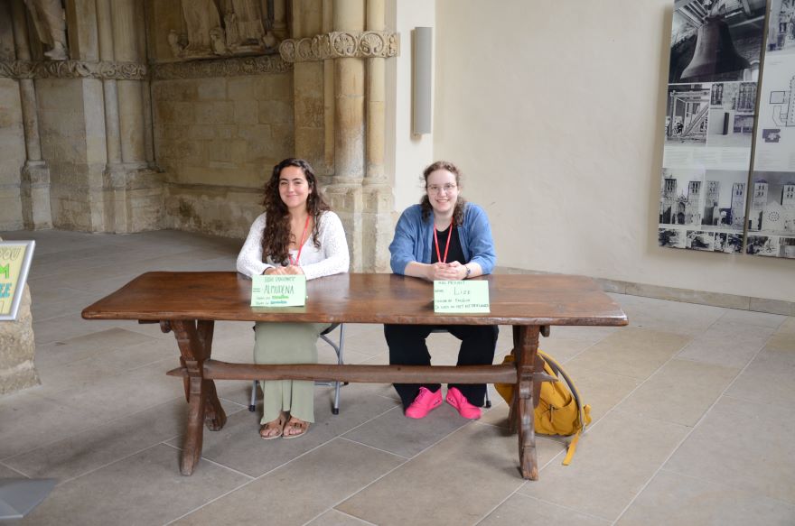 Zwei junge Frauen sitzen hinter dem Paradiesportal des St.-Paulus-Doms hinter einem Holztisch.