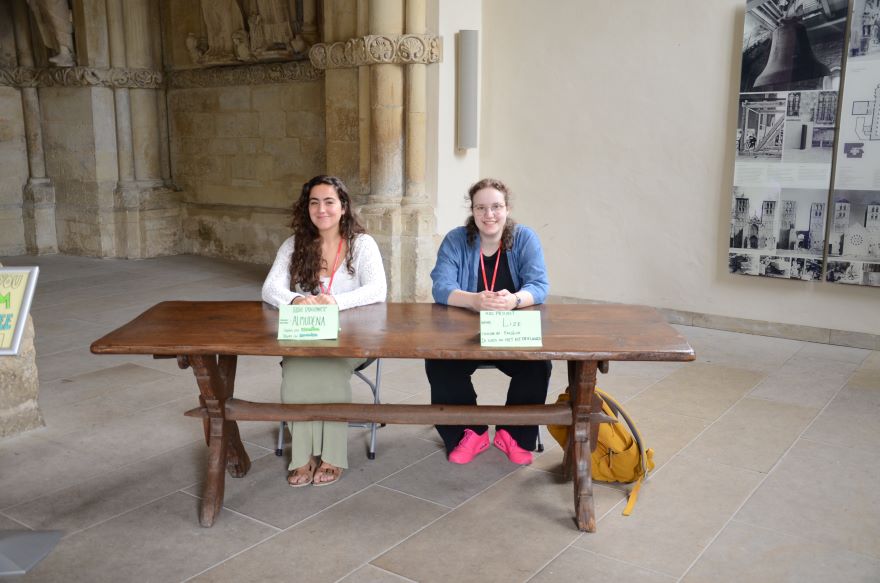 Zwei junge Frauen sitzen hinter dem Paradiesportal des St.-Paulus-Doms hinter einem Holztisch. Zwei junge Frauen sitzen hinter dem Paradiesportal des St.-Paulus-Doms hinter einem Holztisch.