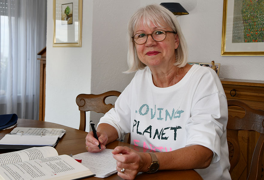 Christa Ressmann sitzt am Tisch und lächelt in die Kamera. Vor ihr liegen verschiedene Unterlagen.