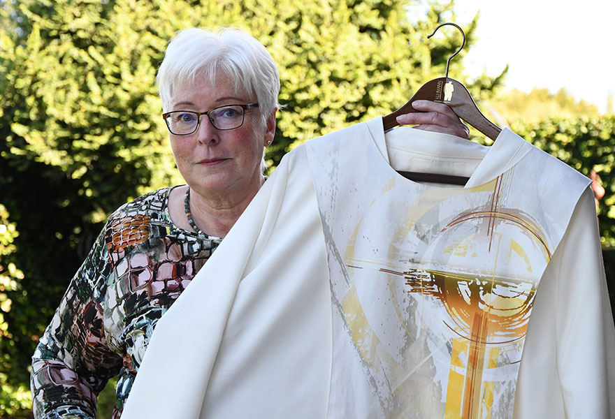 Angelika Surholt hält ein helles Gewand, auf dem in Gelbtönen das Kreuz in einer Sonne aufgeht, in die Kamera. 