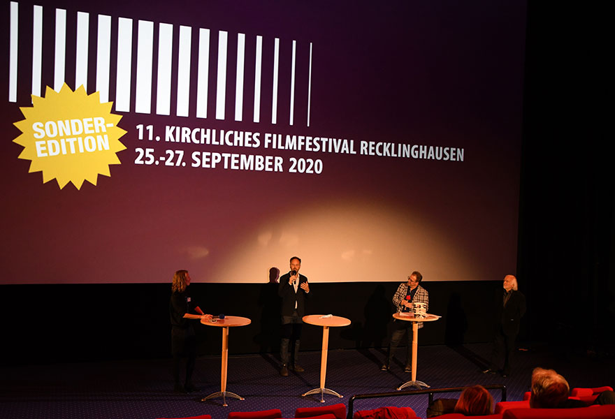 Vor der Leinwand stehen Regisseur Torsten Körner (zweiter von links) den Fragen von Julia Borries (links), Erwachsenenbildungsreferat des evangelischen Kirchenkreises Recklinghausen, sowie der beiden künstlerischen Leiter Horst Walter und Michael M. Kleinschmidt (von rechts) an Tischen. Im Hintergrund ist das Logo des festivals groß zu sehen. .