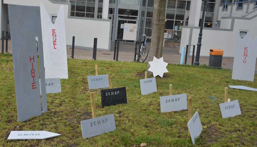 Eine moderne Krippe auf einer Wiese, auf Schildern stehen begriffe wie "Hirte", "Schaf" oder "König".