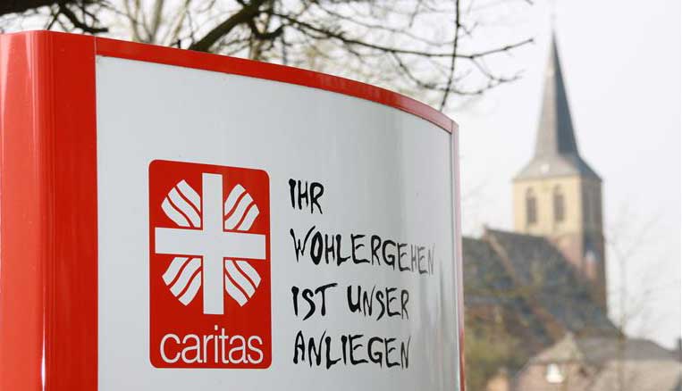 Im Vordergrund ist ein Schild der Caritas zu sehen, im Hintergrund ein Kirchturm.