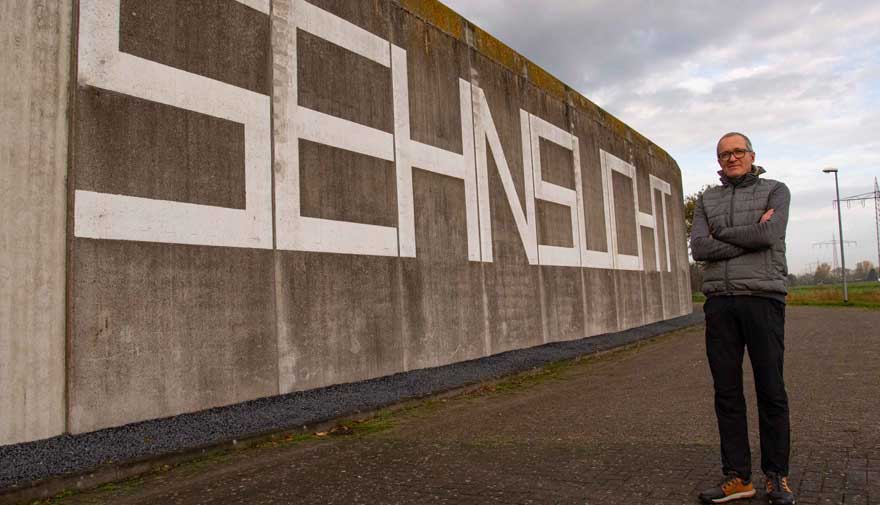 Ein Mann steht vor einer hohen Betonmauer, auf der der Schriftzug "Sehnsucht" zu lesen ist.