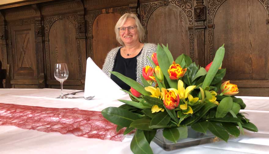 Eine Frau sitzt an einem Tisch, davor steht ein Blumengesteck.