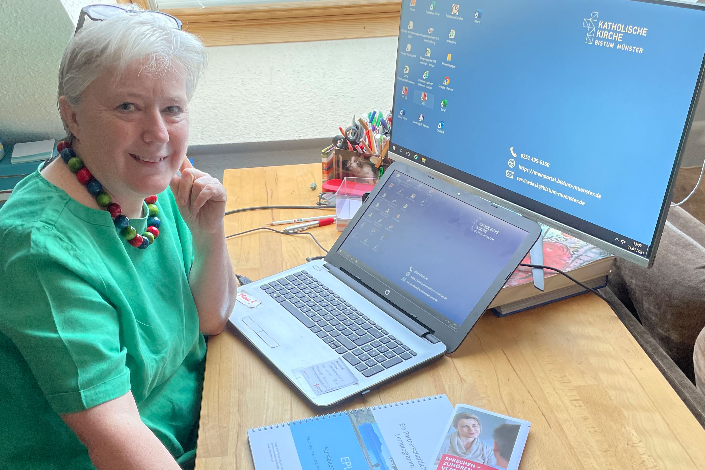 Trainerin Eva Polednitschek-Kowallick sitzt vor einem aufgeklappten Laptop und einem großen Monitor. Auf dem Schreibtisch liegen zwei Broschüren. nikatat Trainerin Eva Polednitschek-Kowallick sitzt vor einem aufgeklappten Laptop und einem großen Monitor. Auf dem Schreibtisch liegen zwei Broschüren. nikatat