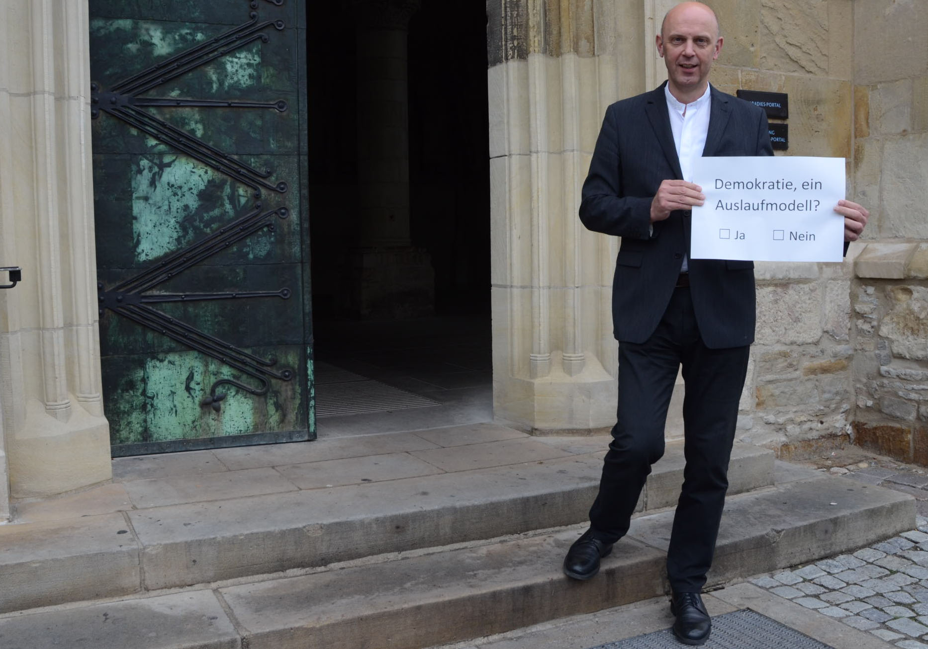 Dompropst Kurt Schulte steht vor der halb geöffneten Eingangstür des Doms. In seinen Händen hält er einen großen weißen Zettel mit der Aufschrift "Demokratie - ein Auslaufmodell?" sowie den Ankreuzmöglichkeiten "Ja" und "Nein".