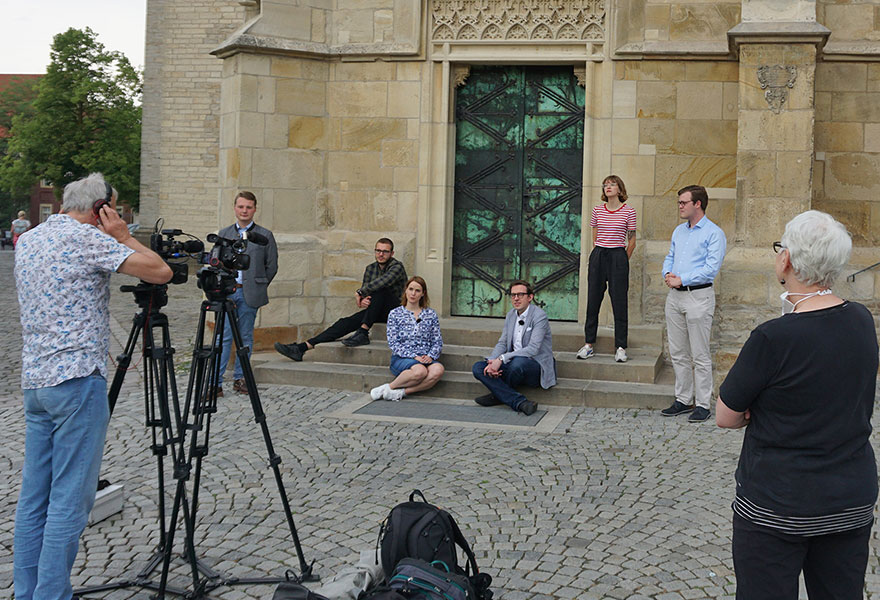 Die Beteiligten sitzen und stehen auf den Stufen vor dem Dom und werden gefilmt.
