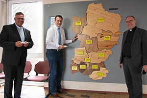 Weihbischof Lohmann steht mit dem Dorstener Bürgermeister Tobias Stockhoff und Pfarrer Dr. Stephan Rüdigern vor einer Karte aus Holz, die die einzelnen Ortsteile Dorstens zeigt.