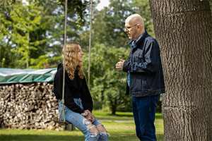 Eine junge Frau Sitzt auf einer an einem Baum hängenden Schaukel. Ein Mann steht vor ihr und spricht zu ihr.