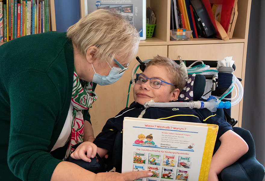 Birgitt Gottfried zeigt ihrem Sohn Paul, der im Rollstuhl sitzt und an Schläuche angeschlossen ist, ein Buch.