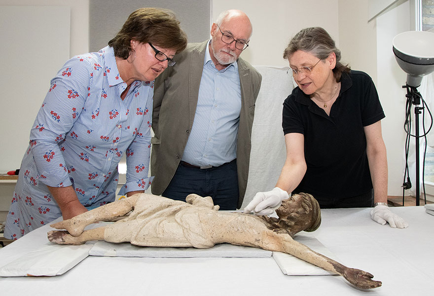 Dr. Martina Dlugaiczyk (links) und Gisbert Stimberg stehen an einem Tisch, auf dem der Korpus liegt. Restauratorin Beate Zumkley erklärt einige Details und zeig mit dem Finger darauf.